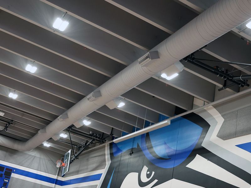 Interior view of a gymnasium ceiling with exposed gray beams, circular ventilation ducts, and bright overhead lights.