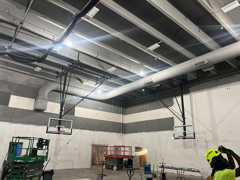 Interior view of a gymnasium construction site with two basketball hoops, exposed ceiling pipes, and mechanical lifts.