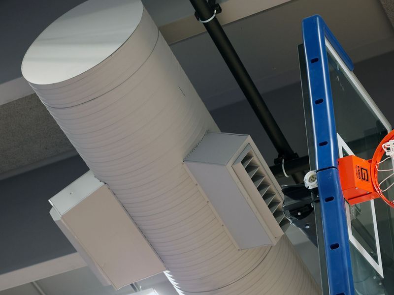 A white cylindrical ventilation duct with square vents installed above a basketball hoop in a gym.