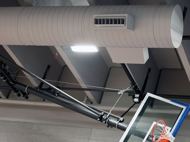 A basketball hoop system is suspended from a gym ceiling near a large ventilation duct and overhead lighting.