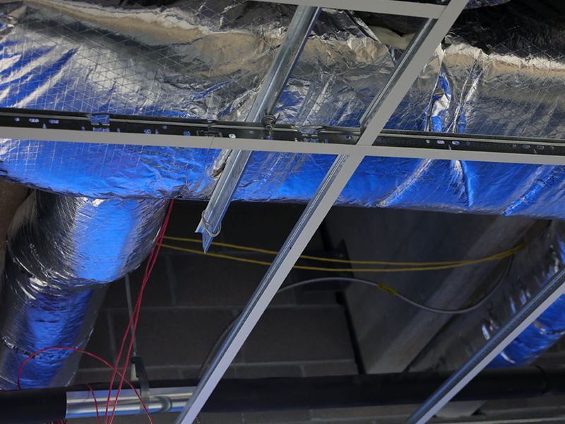 Insulated silver air ducts and exposed wiring installed above a metal grid ceiling frame.
