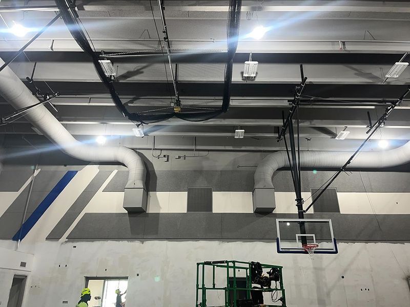 An indoor basketball court with exposed ductwork, gray and white wall panels, a basketball hoop, and a construction lift.