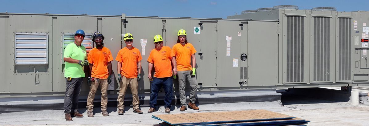 Five people in high-visibility orange and green shirts and hard hats pose in front of a large industrial HVAC unit on a roof.