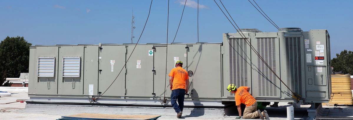 Two workers in high-visibility orange shirts guide a large rooftop HVAC unit being lowered by a crane.