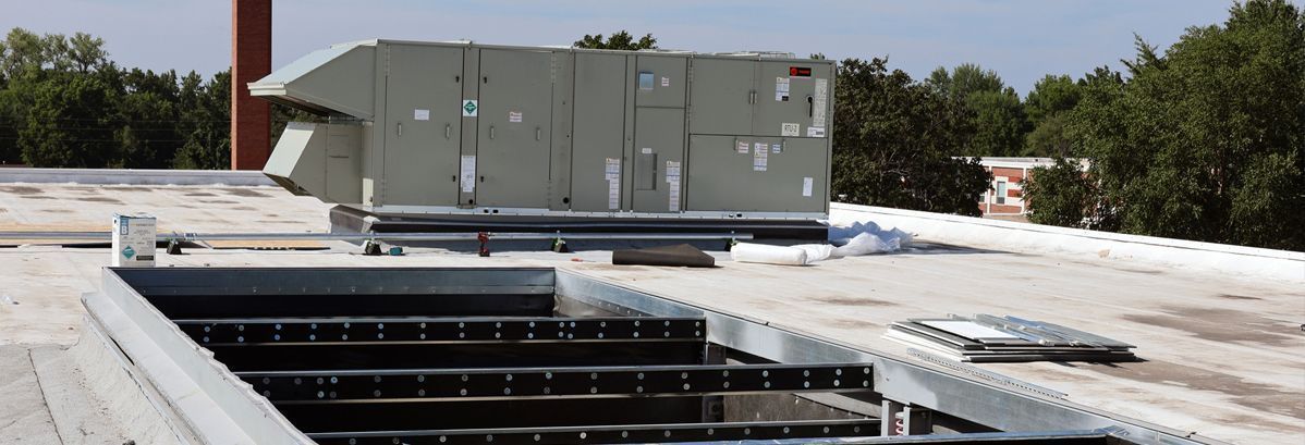 A rooftop scene featuring a large metal HVAC unit next to an open, rectangular roof hatch surrounded by metal flashing.