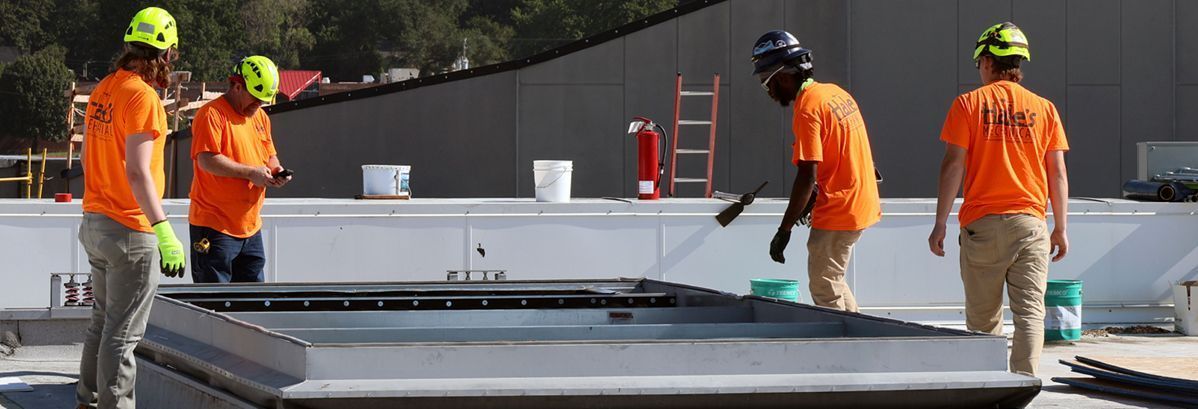 Four people wearing hard hats and orange shirts work on a low, rectangular metal structure on a rooftop.