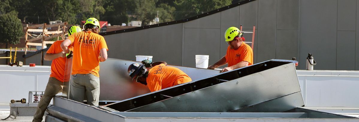 Four construction workers in orange shirts and neon hard hats install metal flashing on a flat commercial roof.