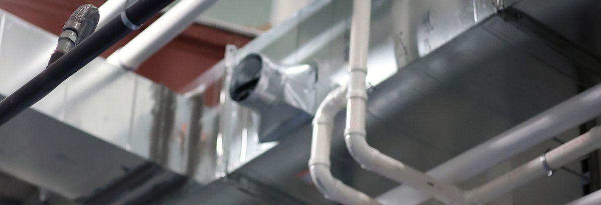 A low-angle view of industrial metal HVAC ductwork and white PVC piping installed overhead in an unfinished building.