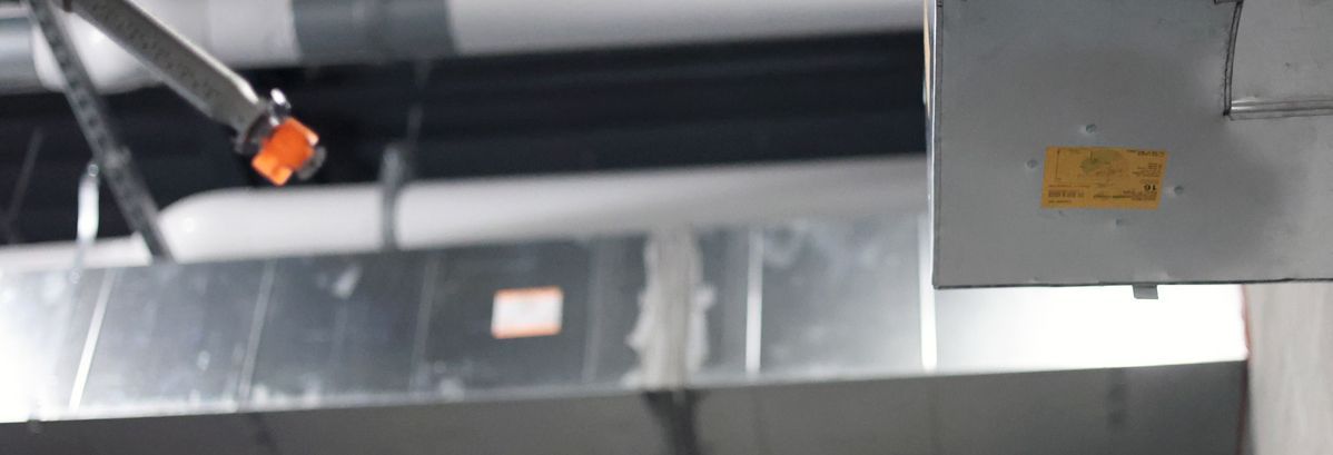 Metal HVAC ducts and a yellow warning label hang from a ceiling in an industrial or commercial building.