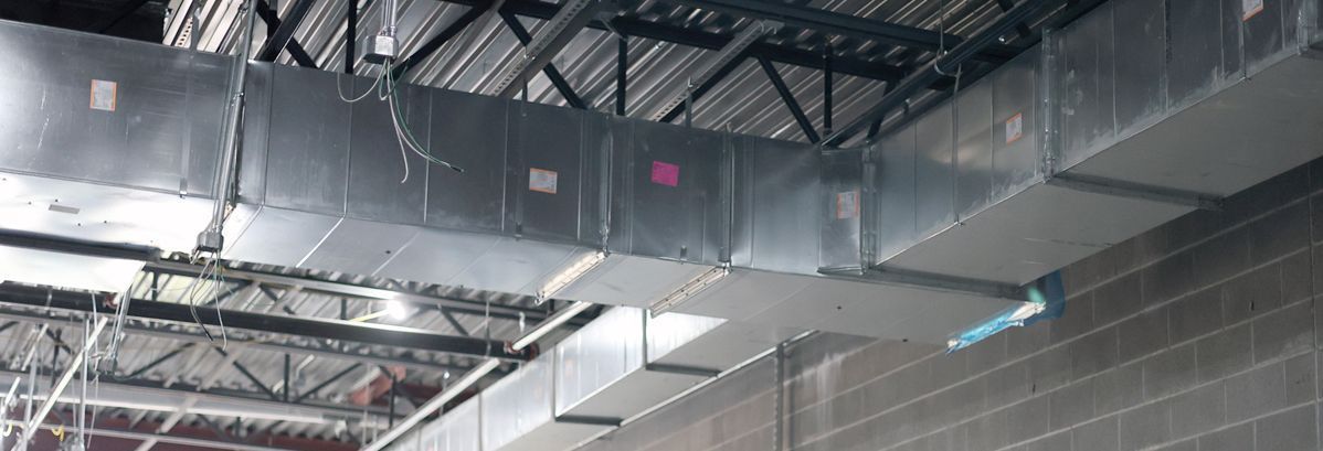 Large metal HVAC ductwork runs horizontally along the ceiling of a building with exposed rafters and a concrete wall.
