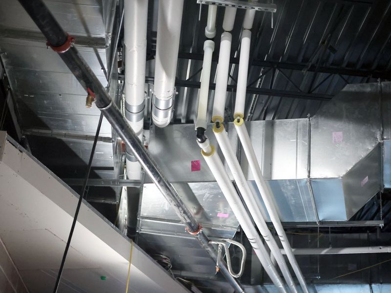 Industrial ceiling view featuring complex metal ductwork, suspended piping, and conduit against a dark, exposed structure.