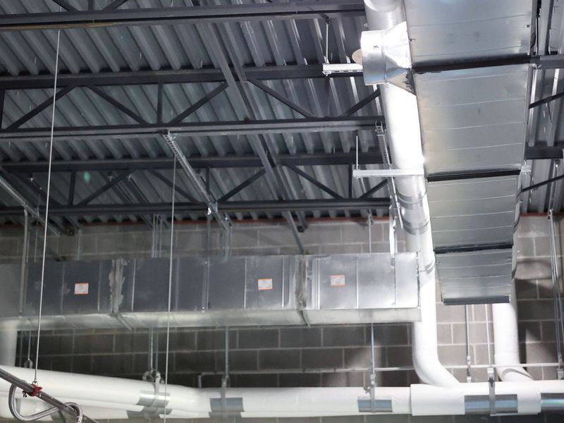 Exposed ductwork and pipes hang from a metal ceiling structure in an industrial building with concrete block walls.