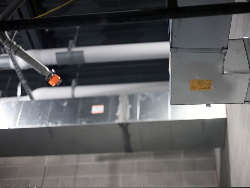 Industrial ceiling pipes and metal HVAC ducts exposed above a concrete block wall in an unfinished interior space.