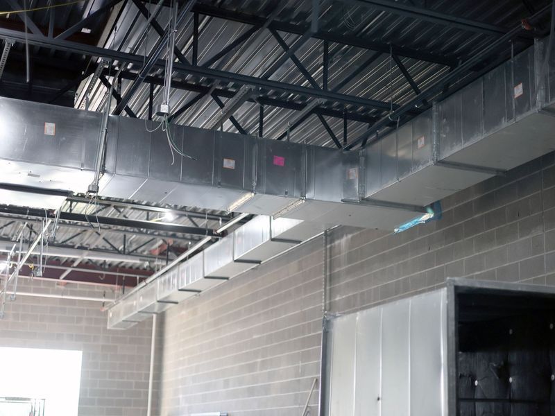 A view of industrial HVAC metal ductwork suspended from a dark ceiling above a concrete block wall in a commercial space.