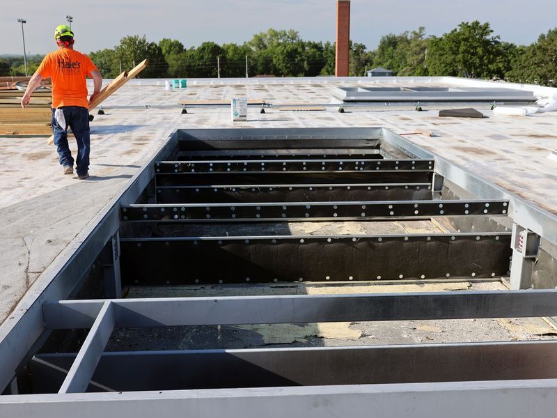 A person in a bright orange shirt walks across a flat rooftop toward a large rectangular opening in the building structure.