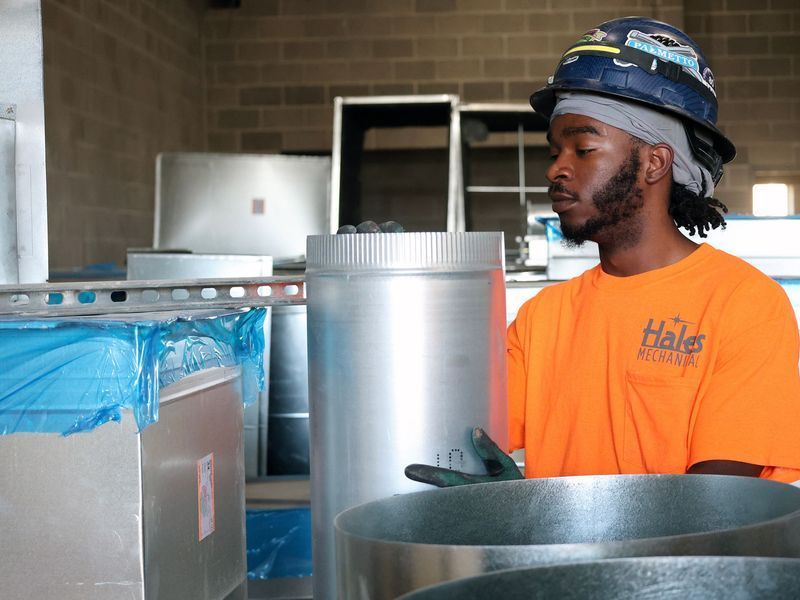 A worker in an orange shirt and hard hat holds a piece of cylindrical metal ductwork in a workshop.