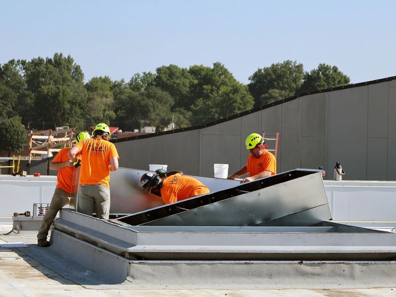 Construction workers in orange shirts and hard hats working on a metal rooftop installation under a clear blue sky.