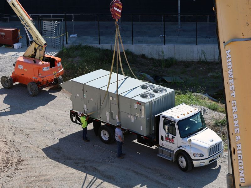 A crane lifts a large industrial HVAC unit from a flatbed truck while two workers stand nearby on a dirt lot.