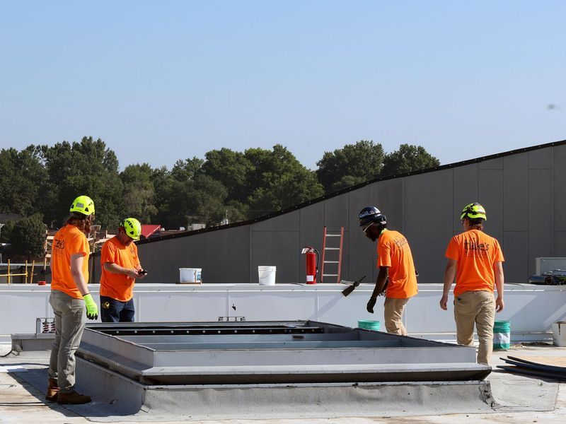 Four workers in orange shirts and hard hats work on a flat roof around a rectangular skylight frame on a sunny day.