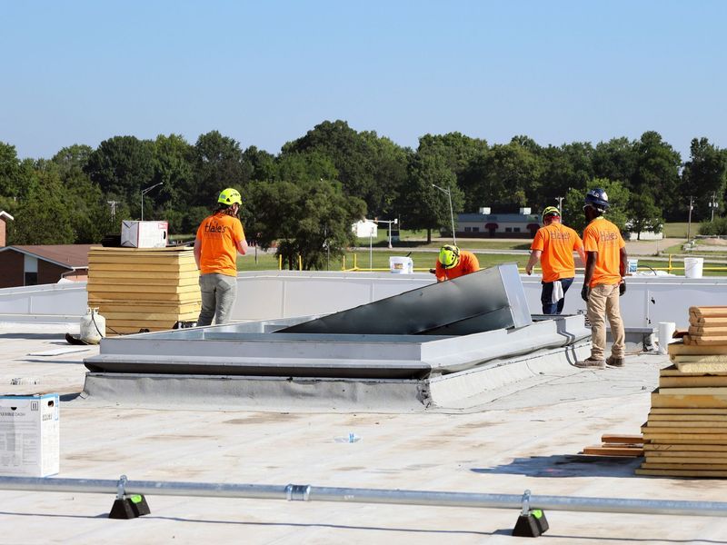 Construction workers in orange shirts work on a flat roof, installing or repairing a skylight on a sunny day.