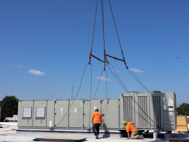 Two workers in orange shirts guide a large HVAC unit being lowered by a crane onto a rooftop under a clear blue sky.