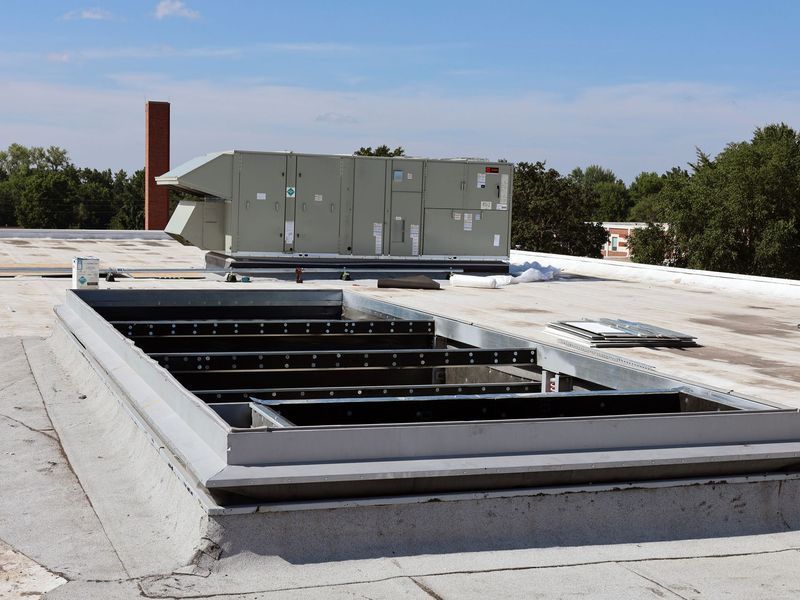 A roof with a large, open metal curb frame in the foreground and an HVAC unit in the background under a blue sky.