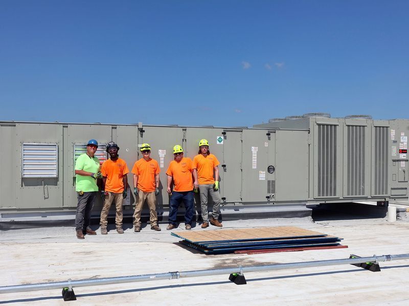 Five people in work uniforms and safety gear stand in a line on a rooftop next to a large HVAC industrial unit.