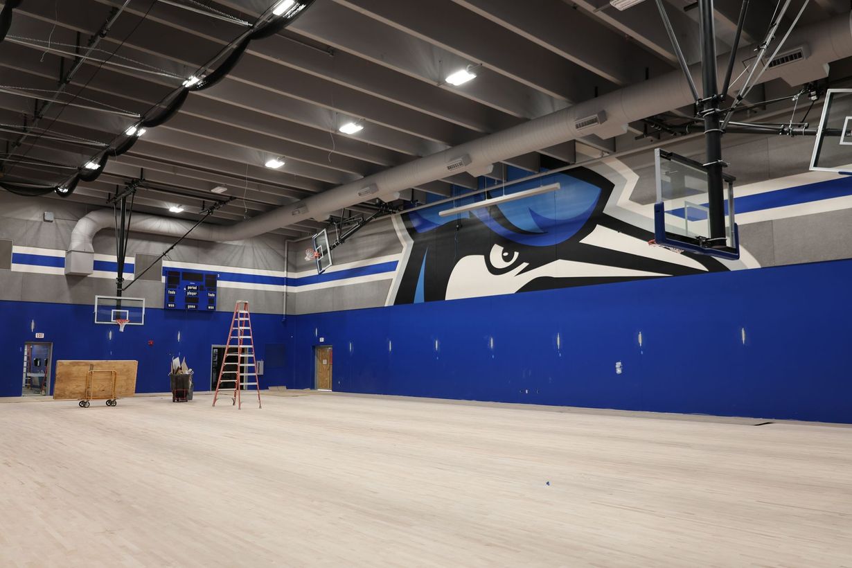 An indoor basketball court with a blue wall featuring a large bird mascot mural, a ladder, and a scoreboard.