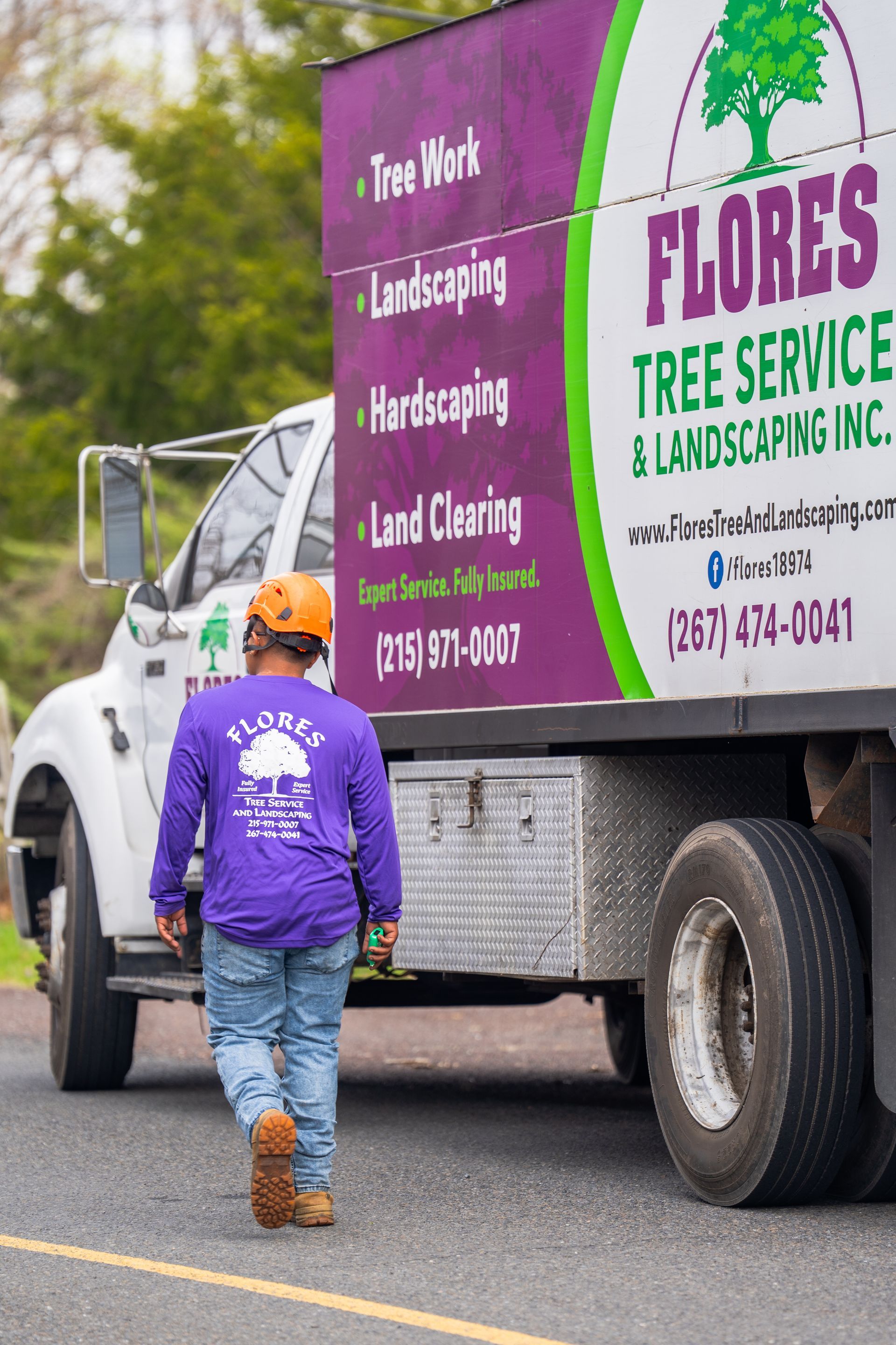 Worker in orange hard hat and purple shirt beside a Flores Tree Service landscaping truck