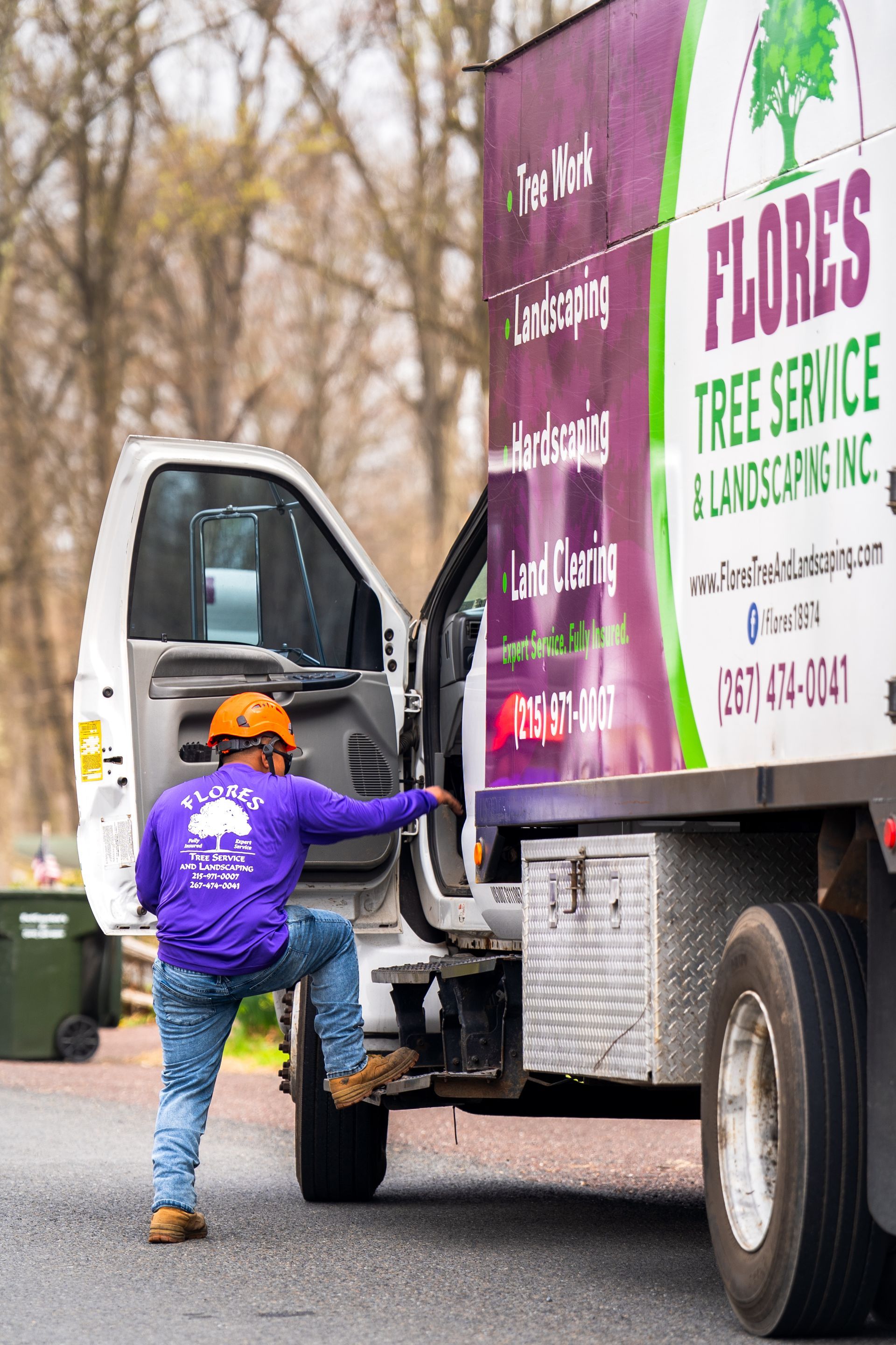 Worker in purple hoodie steps onto a truck beside a green-and-white tree service sign.