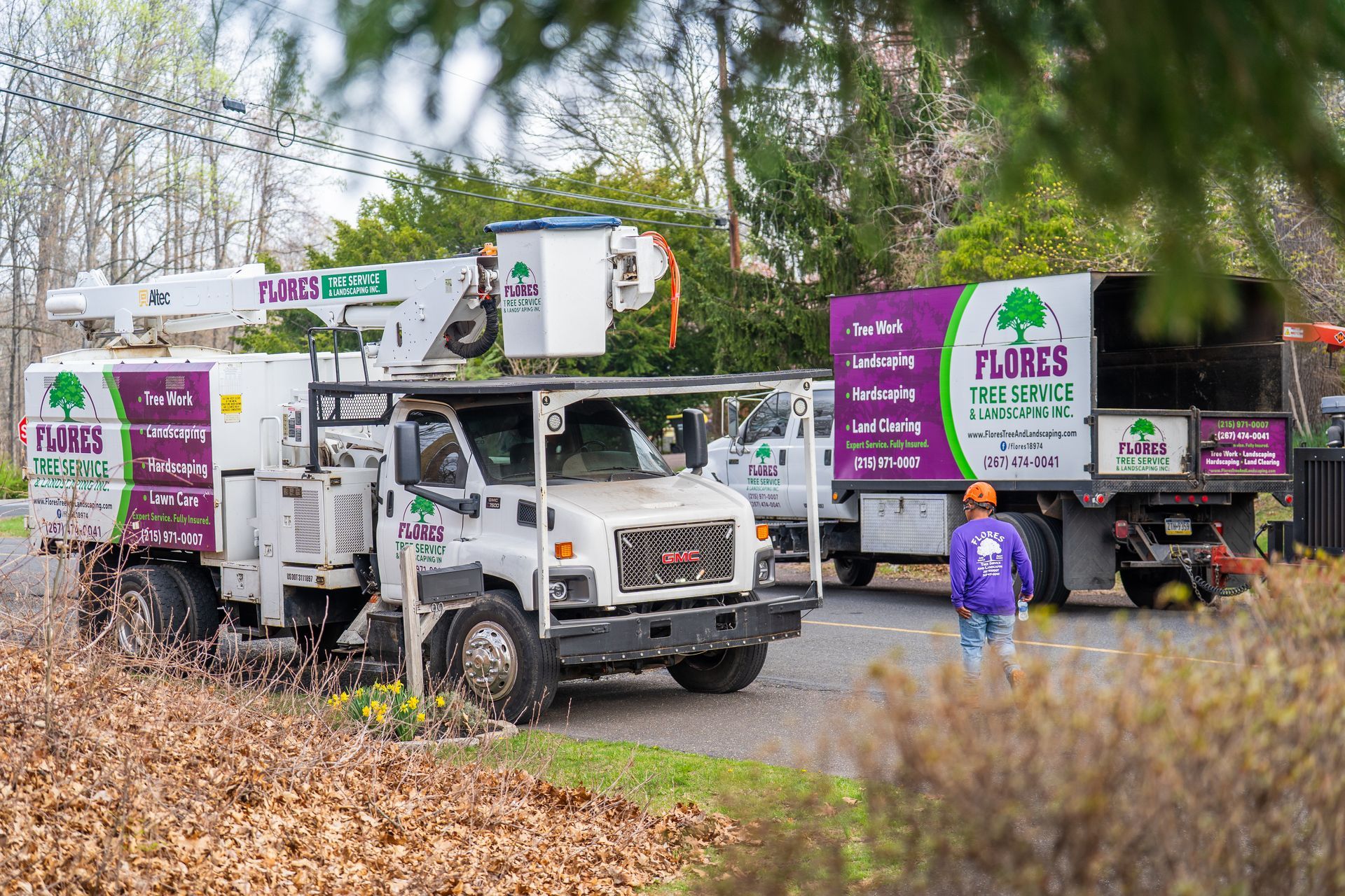 Two white and purple utility trucks in a leafy outdoor area, likely tree service vehicles.