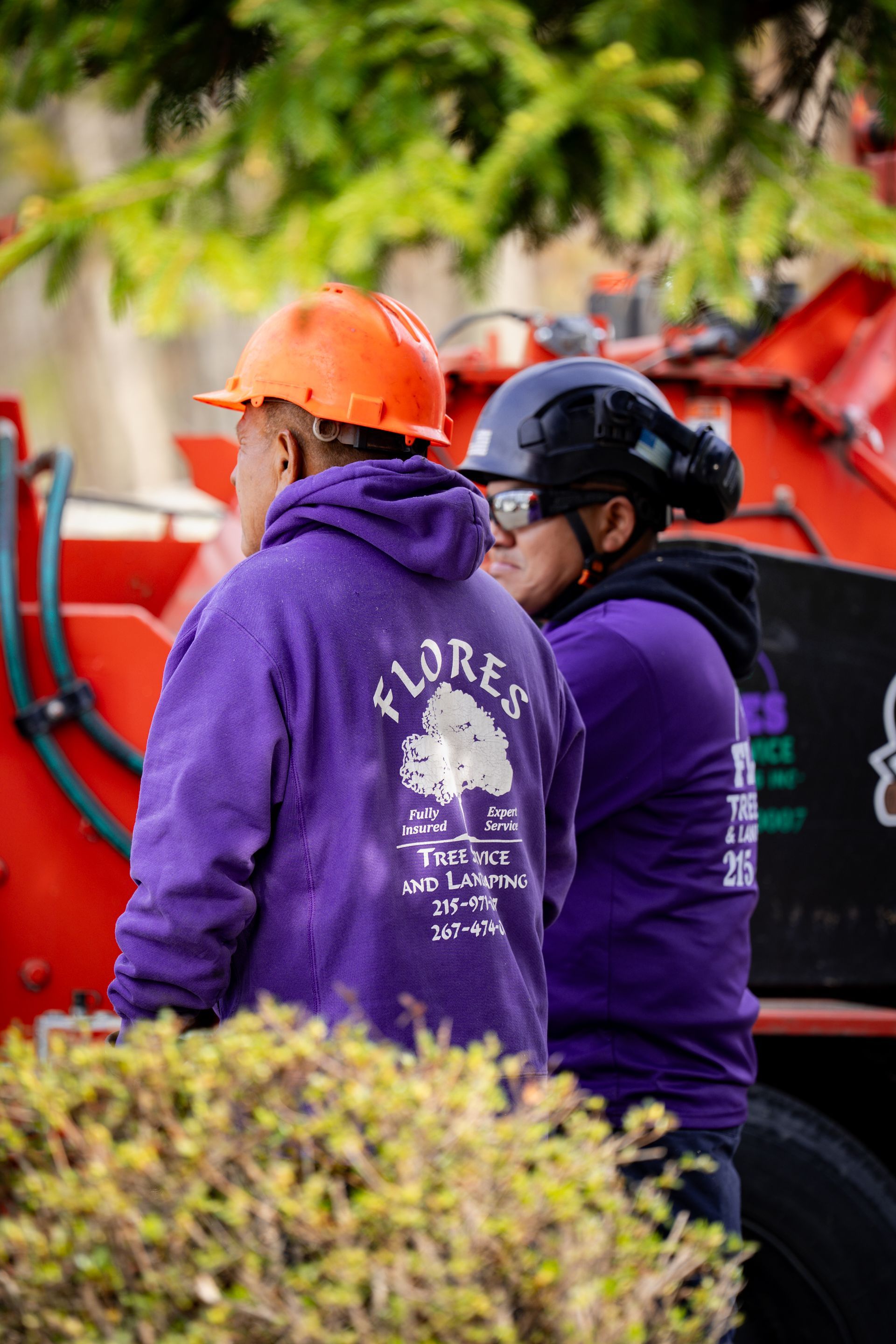 Two workers in purple hoodies and hard hats stand beside a red machine outdoors.