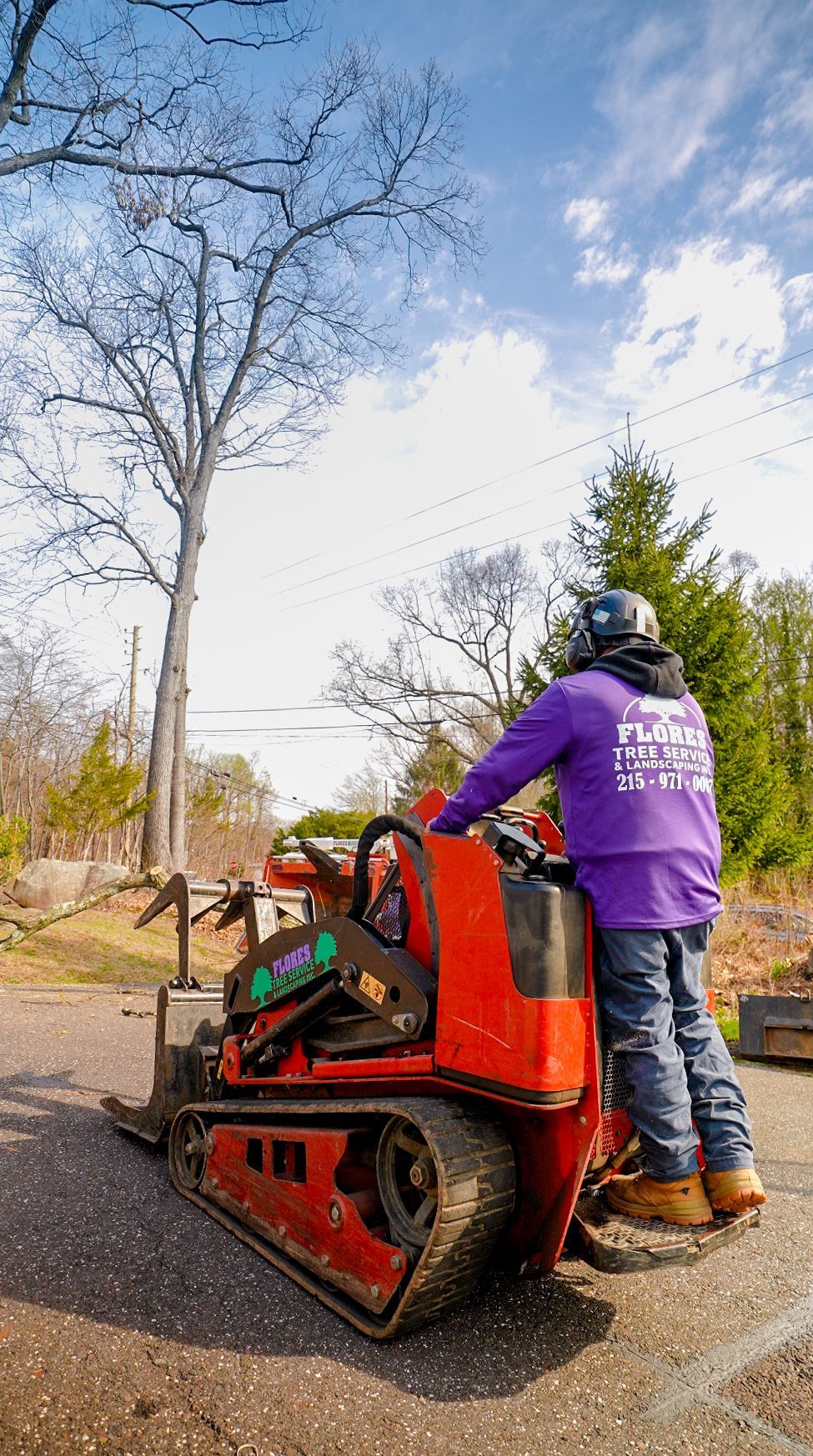 Worker operating an orange tracked machine on a gravel path in a wooded area, wearing a purple jacket and helmet