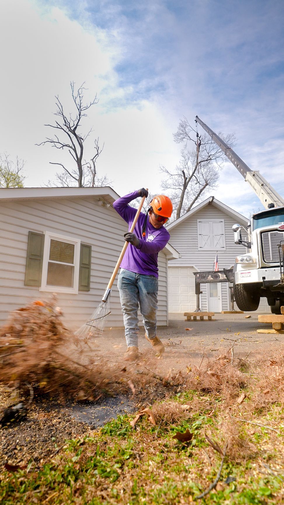 Worker in orange hard hat raking debris in a yard beside a truck and crane.