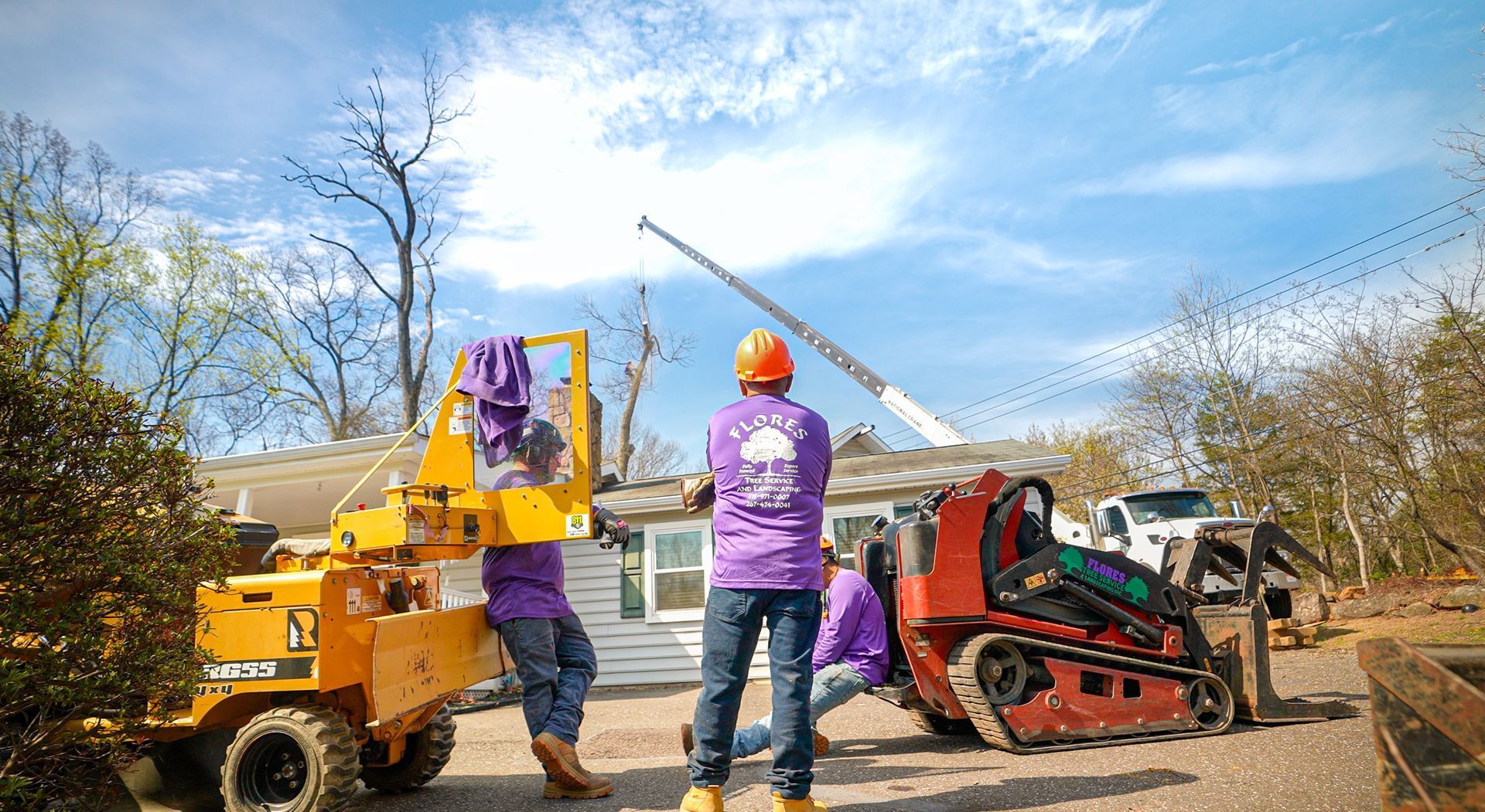 Workers in purple shirts operate a yellow wood chipper beside a red tractor under a blue sky.