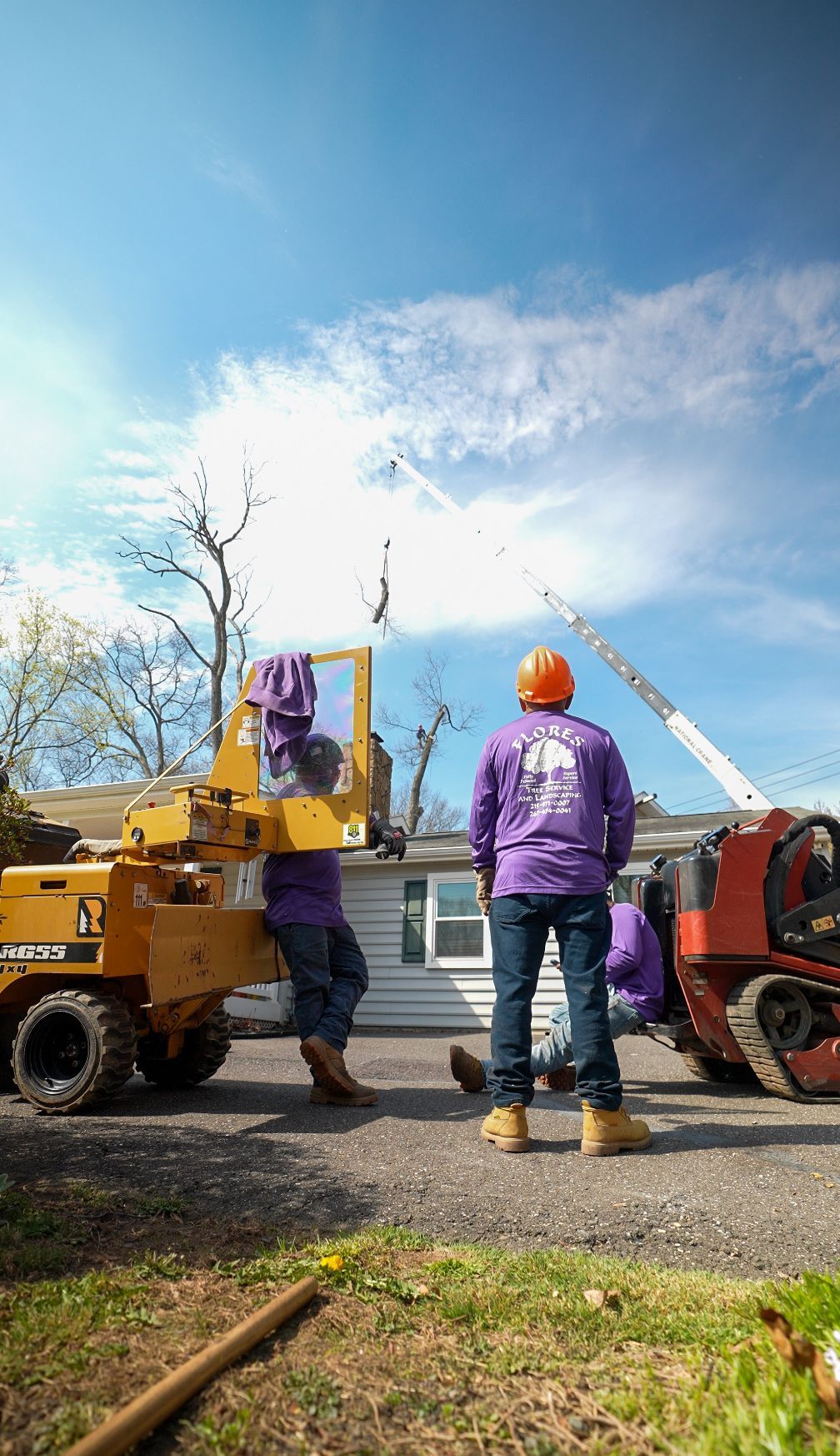 Two workers in purple shirts by a yellow boom lift, with a white bucket truck and crane under a blue sky.