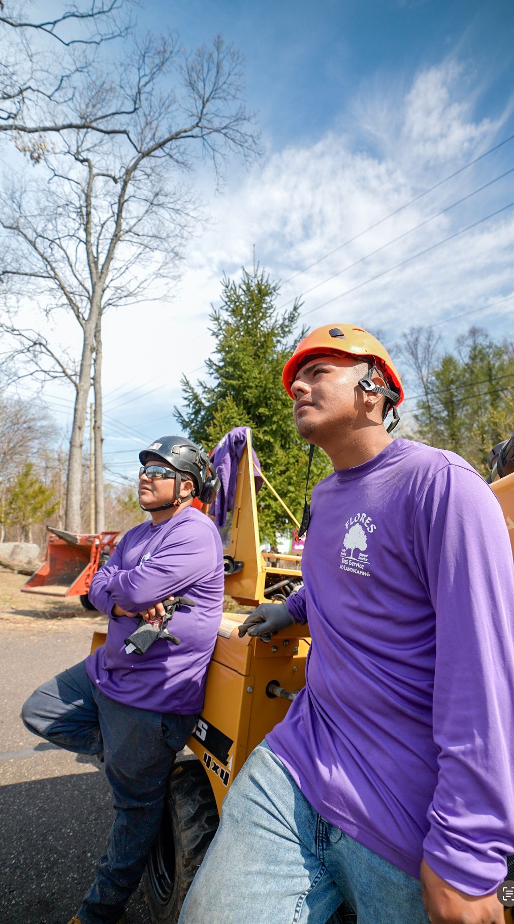 Two people in purple shirts and hard hats stand outdoors near construction equipment under a blue sky.