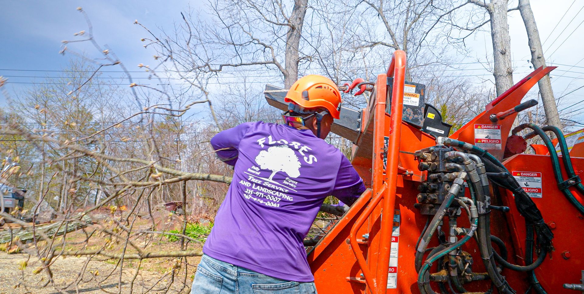 Worker in orange hard hat and purple shirt operating a red wood chipper outdoors.