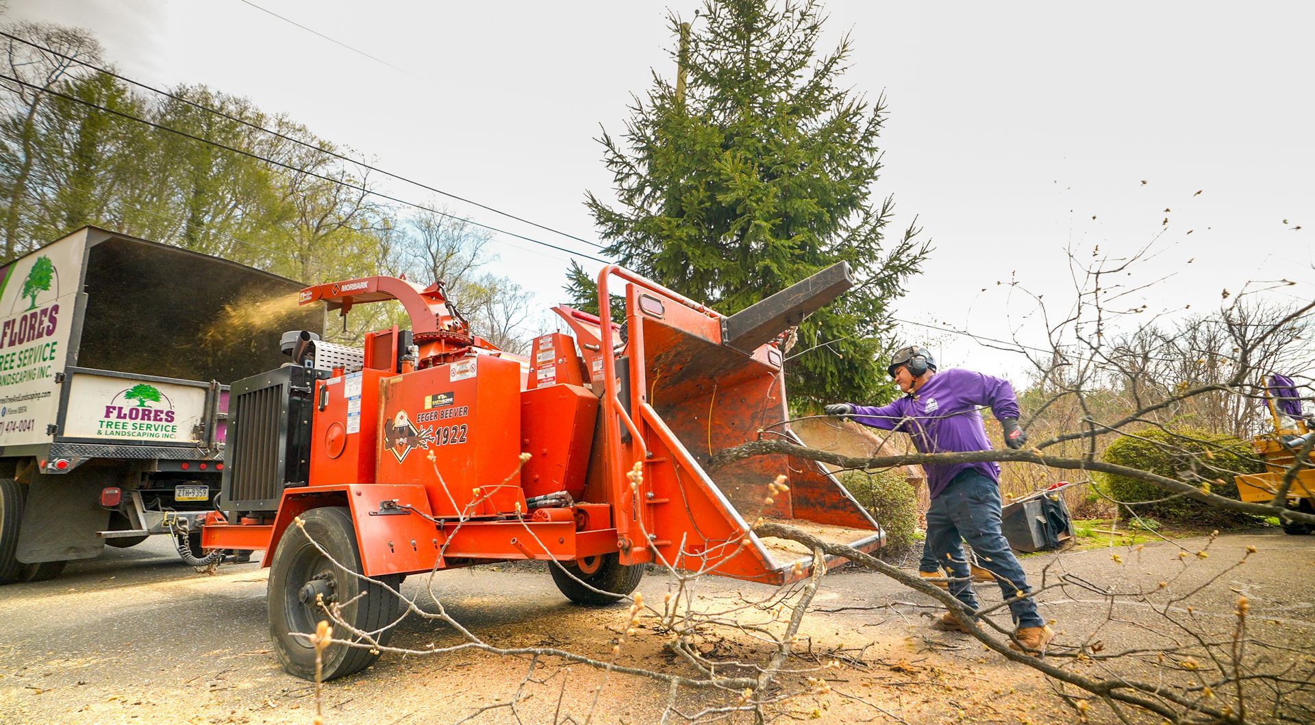 Worker in purple using a wood chipper to shred branches beside a truck on a leaf-covered street
