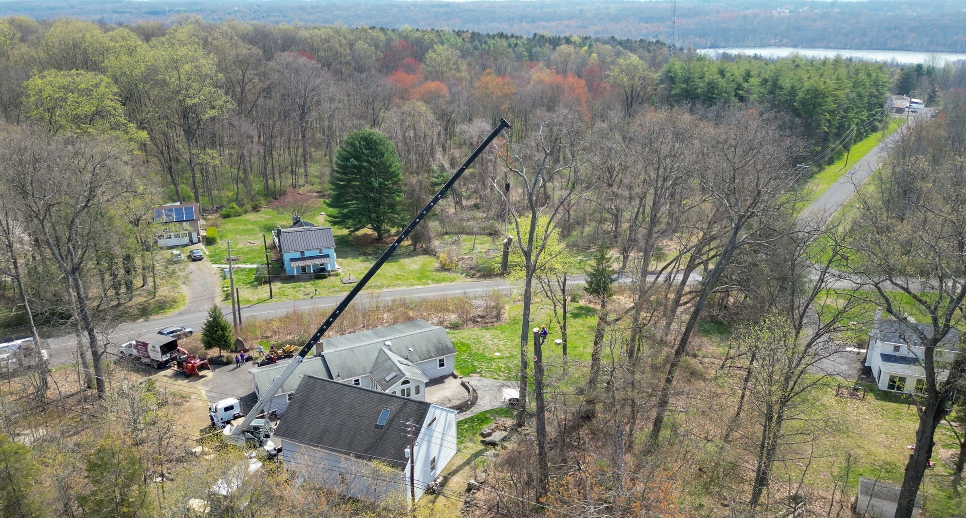 Aerial view of a wooded rural property with houses, roads, and a crane lifting near a roof.