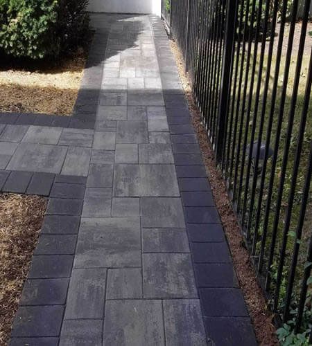 Paver walkway with dark border next to black fence and brown mulch, leading to a white door.