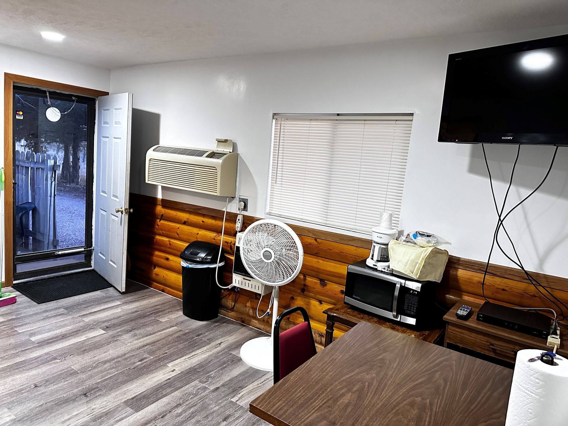 A room with a table , chairs , a fan , a microwave and a flat screen tv.
