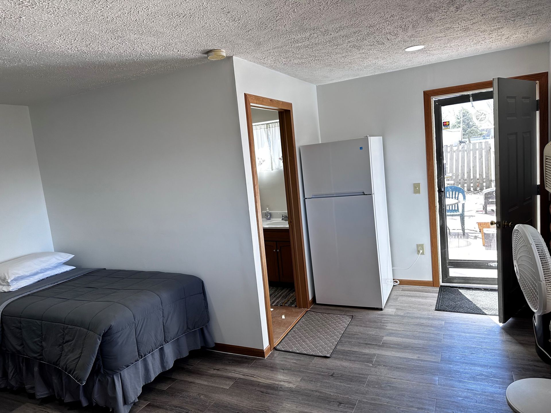 A bedroom with a bed , refrigerator , and a fan.