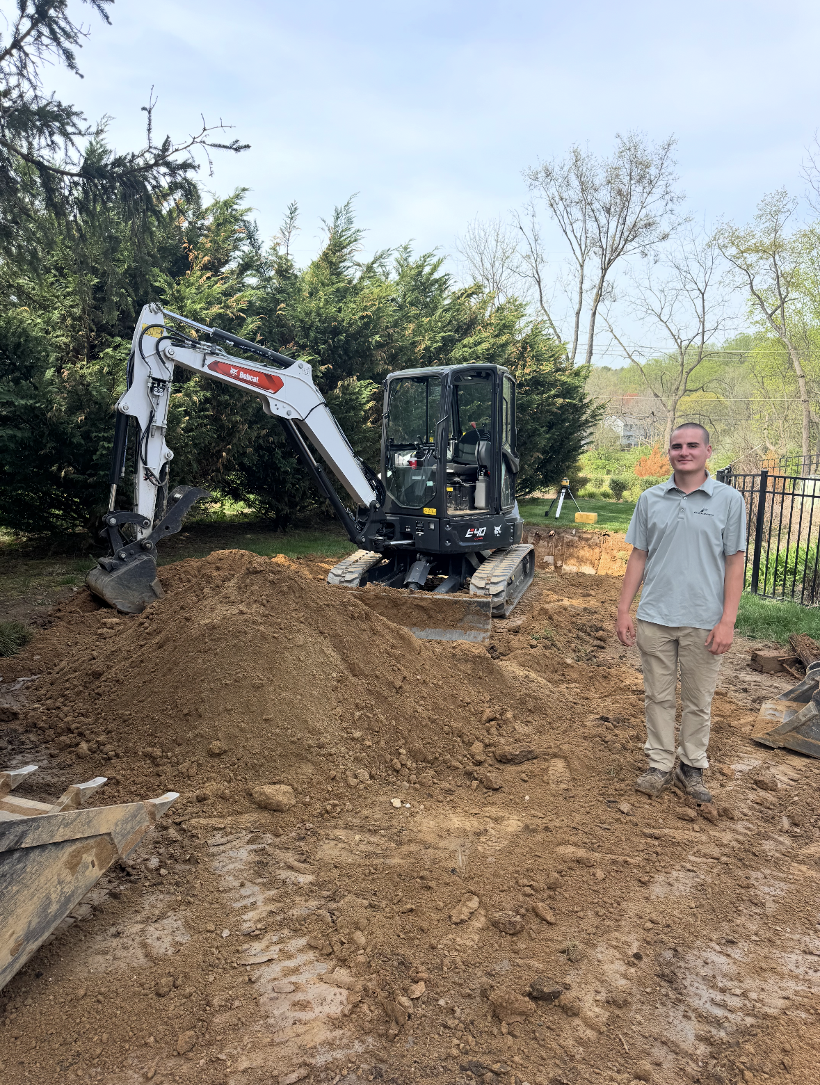 Small excavator on dirt pile beside a smiling man in a yard with trees and a fence.
