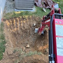 A red excavator arm digging a hole in the ground near a residential air conditioning unit.