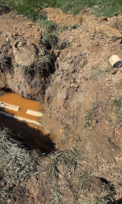 A rectangular trench filled with muddy water, exposing a light-colored pipe, set in an area of patchy dirt and grass.