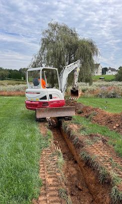 A white and red mini-excavator digs a trench in a grassy field in front of a large weeping willow tree.