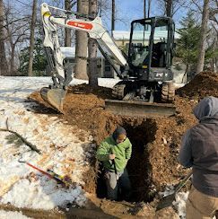 A person stands in a deep trench being dug by a white Bobcat excavator in a snowy, wooded yard.