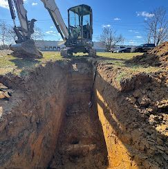 An excavator parked beside a deep, narrow rectangular trench cut into brown earth on a sunny day.
