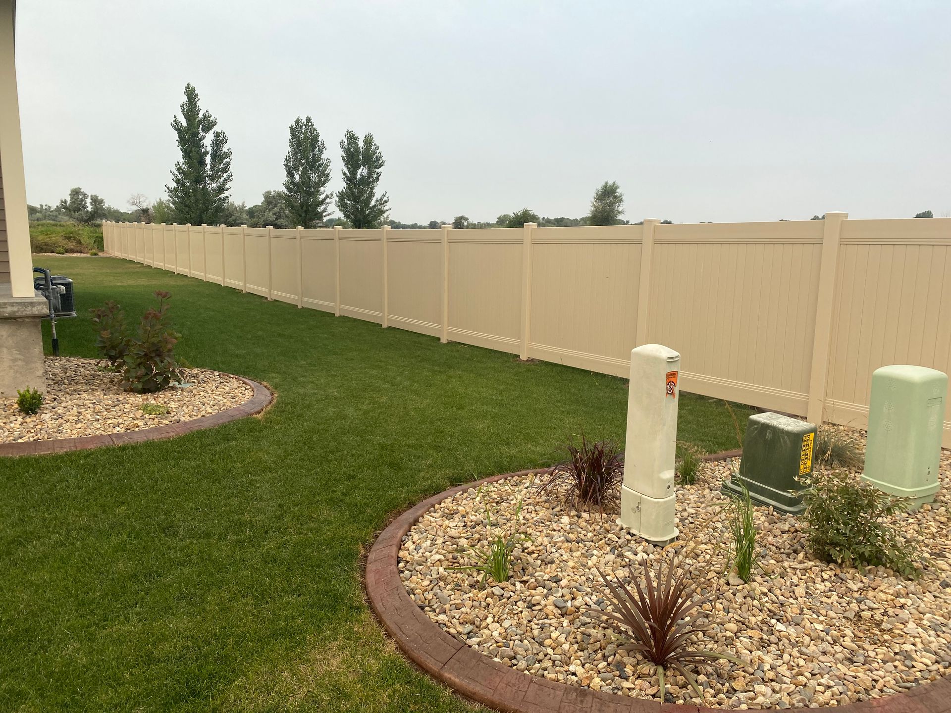 A long beige vinyl fence in a backyard, with green grass, landscaping, and a cloudy sky.